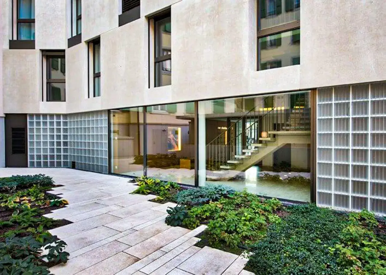 Cortile moderno con pavimentazione in pietra, aiuole verdi e una grande vetrata che rivela una scala interna. Pareti laterali con blocchi di vetrocemento, forse per un'autorizzazione edilizia.