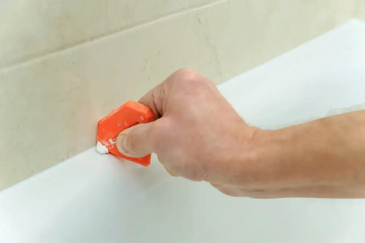 Mano usa spatola arancione per rimuovere silicone da bordo vasca bianca, vicino a piastrelle beige.