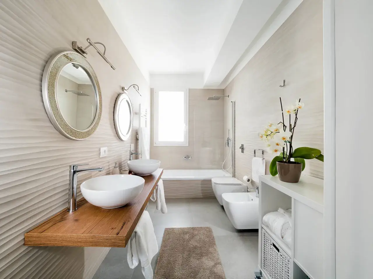 Bagno moderno con doppio lavabo, bidet, wc e vasca sotto finestra. Orchidee bianche decorano il mobiletto.